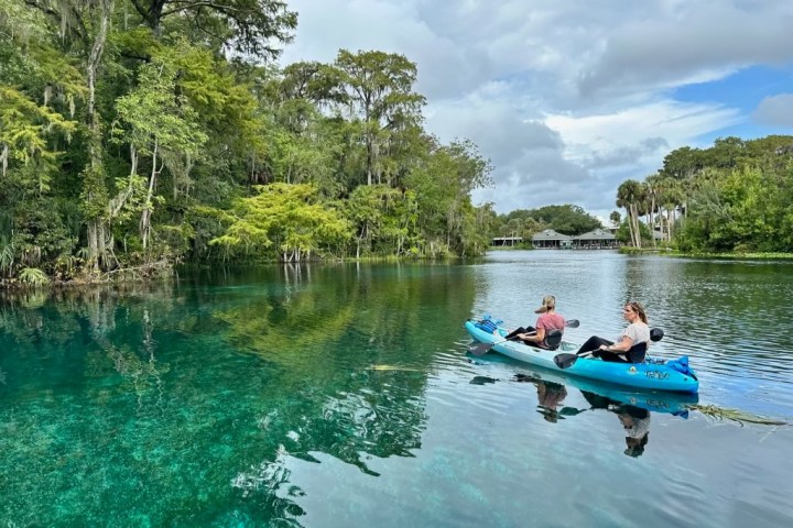 Two people kayaking on a clear, calm river surrounded by lush, green trees.