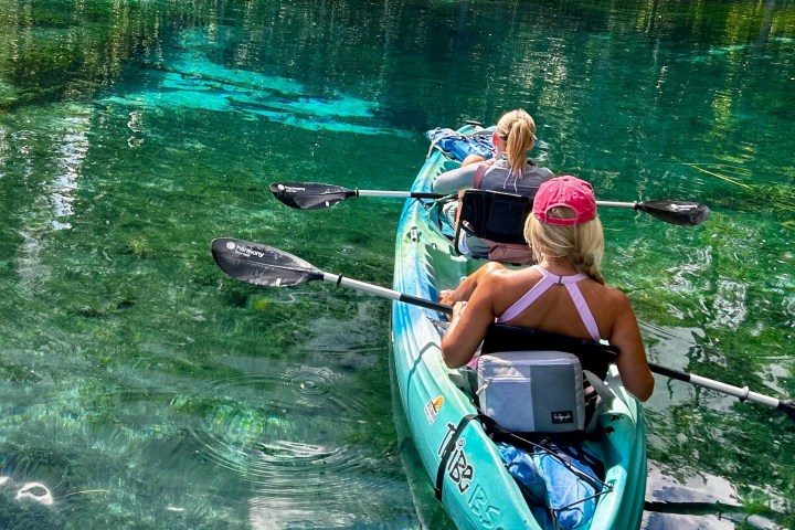 Two people kayaking on clear, turquoise water with lush greenery around.