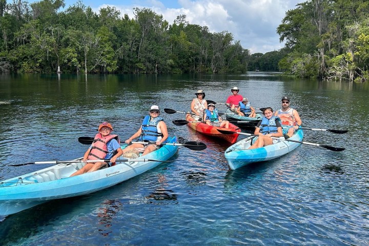 Group kayaking on a calm river surrounded by trees under a cloudy sky.