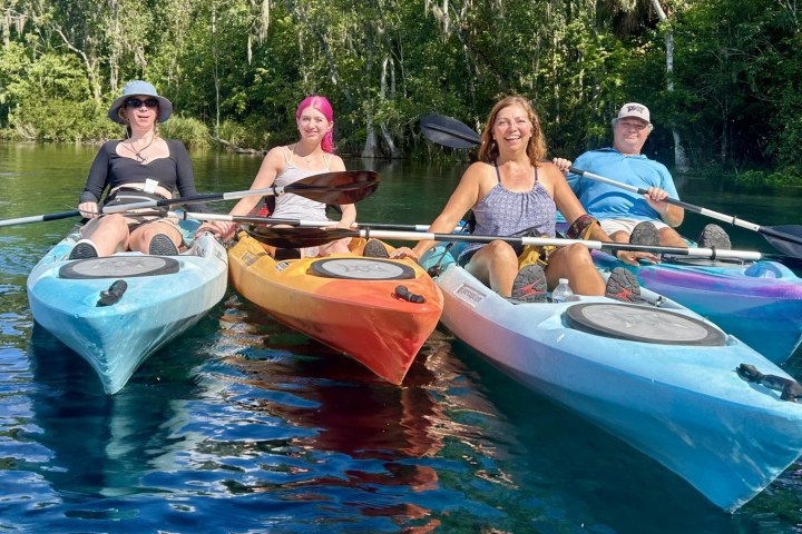 Four people kayaking on a calm river with green trees in the background.