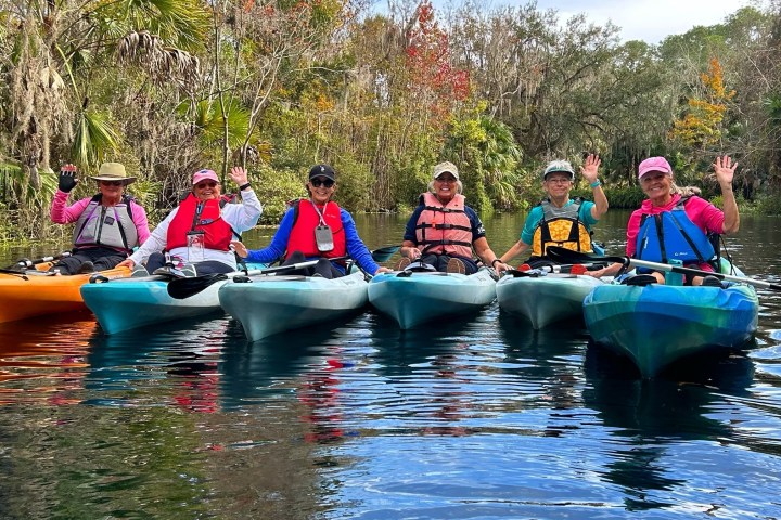 Six people in kayaks on a river surrounded by trees, waving and smiling.