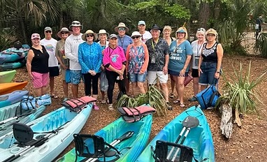 Group of people standing behind kayaks in a wooded area, wearing casual outdoor clothing and hats.